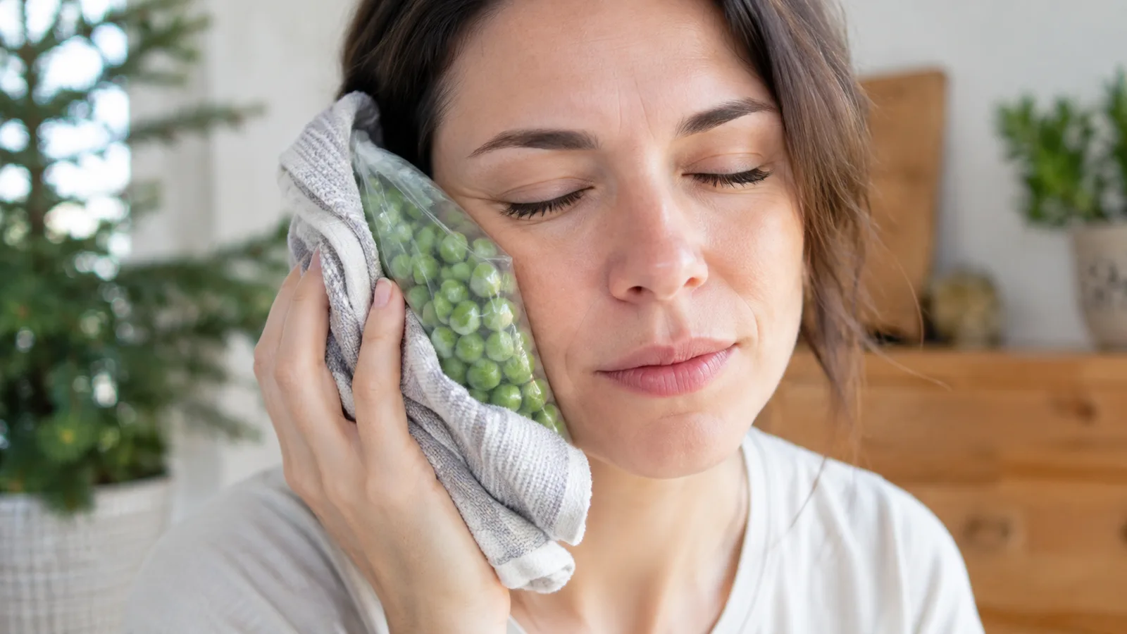 A woman with her eyes closed holds a cloth-wrapped bag of frozen peas against her cheek.